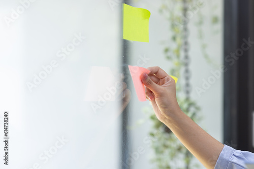 Young Creative business asian woman writing on sticky notes on a glass wall, female colleague looking.