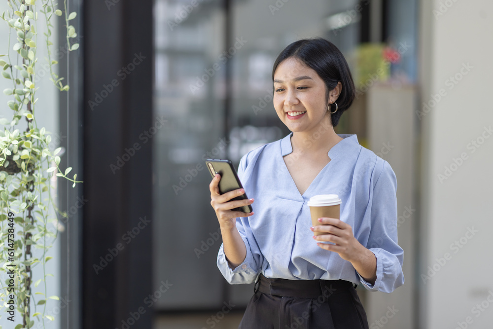 Young beautiful Asian woman holding coffee paper cup and looking at smartphone while sitting at workplace. Happy university student girl using mobile phone. Business woman drinking coffee and smiling. Young beautiful Asian woman holding coffee paper cup and looking at smartphone while sitting at workplace. Happy university student girl using mobile phone. Business woman drinking coffee and smiling.