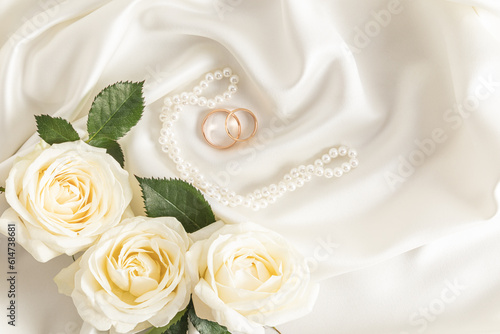 Two gold wedding rings on a delicate satin fabric background with pearl beads and white roses. A copy of the space. Design layout.