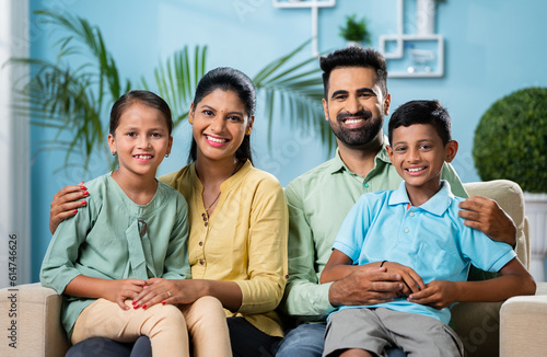 Happy smiling indian couple with sibling kids looking or posing to camera while sitting on sofa at home - concept of nuclaer family, parenthood and generation. © WESTOCK