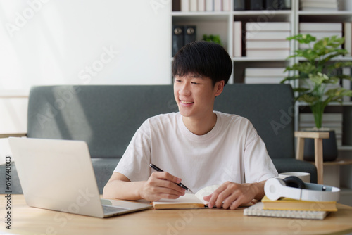 Online education, e-learning. Asian man in stylish casual clothes, studying using a laptop, listening to online lecture, taking notes, online study at home