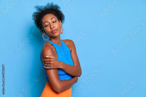 Close up of pretty black girl in glasses, stands in beautiful pose against blue wall, hugging herself closing her eyes, wears blue top, relax concept, copy space