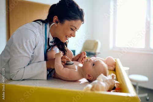 Happy female pediatrician examining baby with stethoscope at doctor's office.