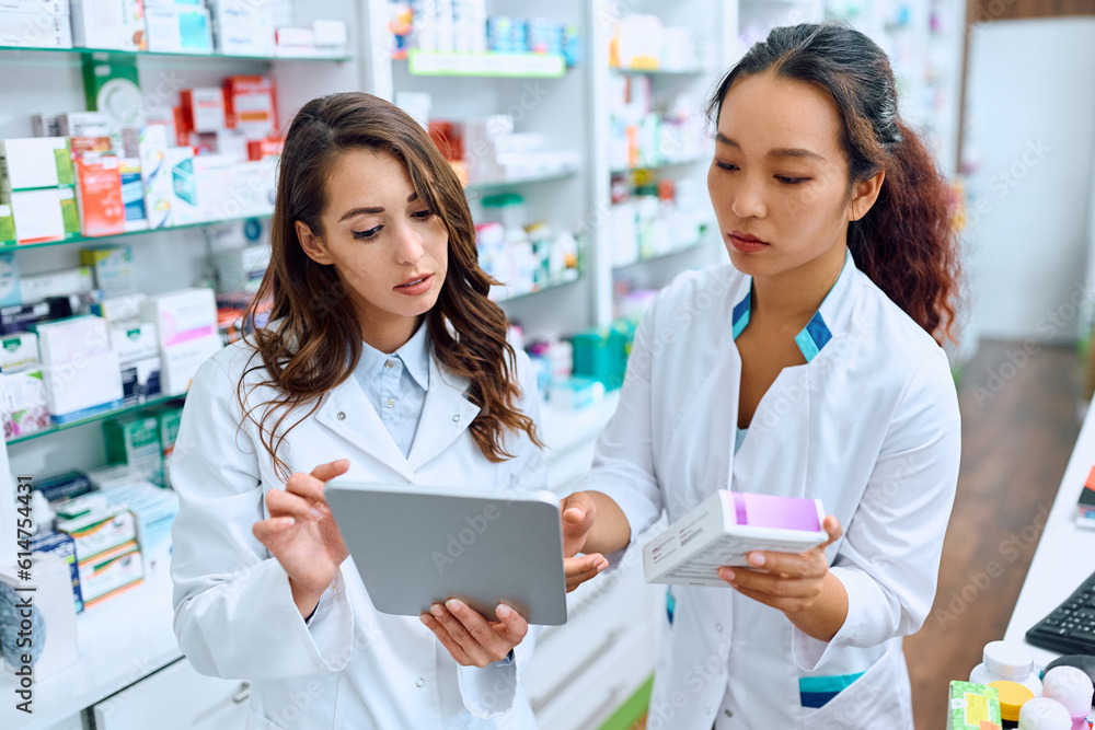 Female pharmacists cooperate while using touchpad and working in drugstore.