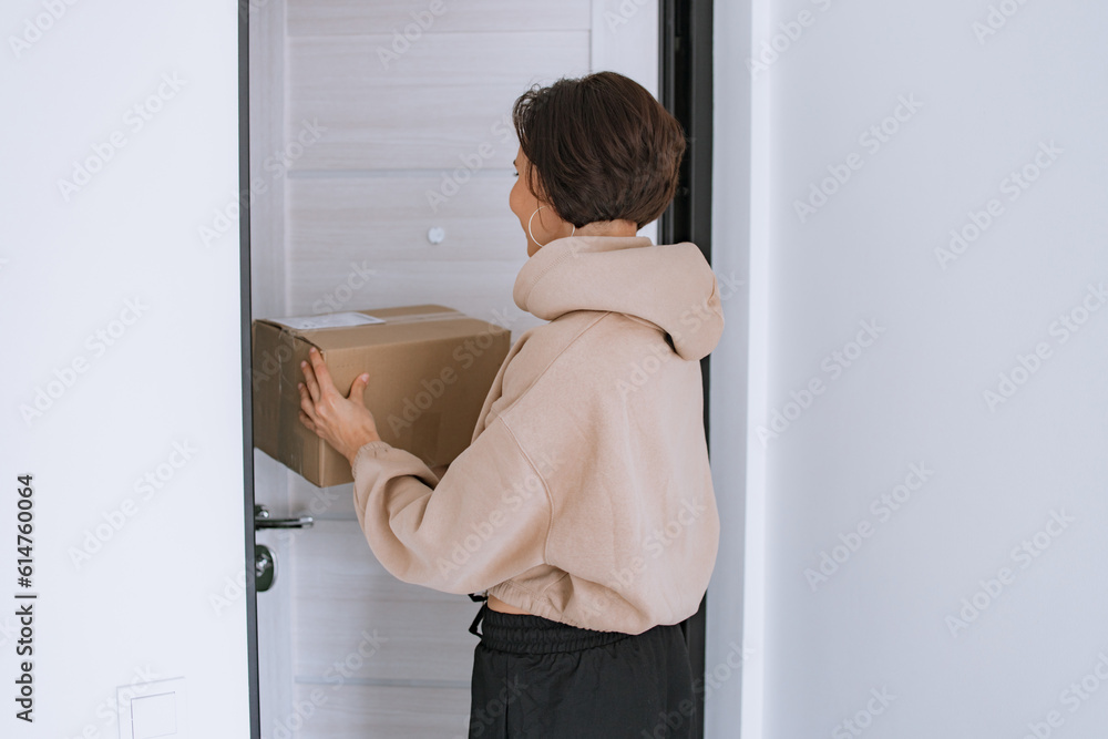 A woman accepts a parcel, a cardboard box with a white label standing at home opening the door. Post delivery