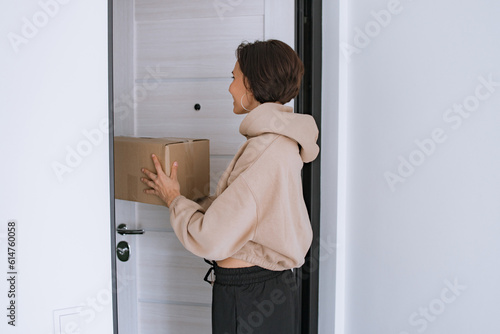 A woman accepts a parcel, a cardboard box with a white label standing at home opening the door. Post delivery