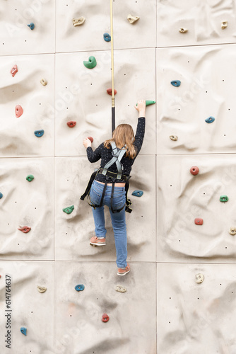 Teenage teen girl grabbing artificial high climbing wall with split ledges. Climbing harness safety sports equipment. Rope amusement city park, obstacle course. Kids children's attraction center
