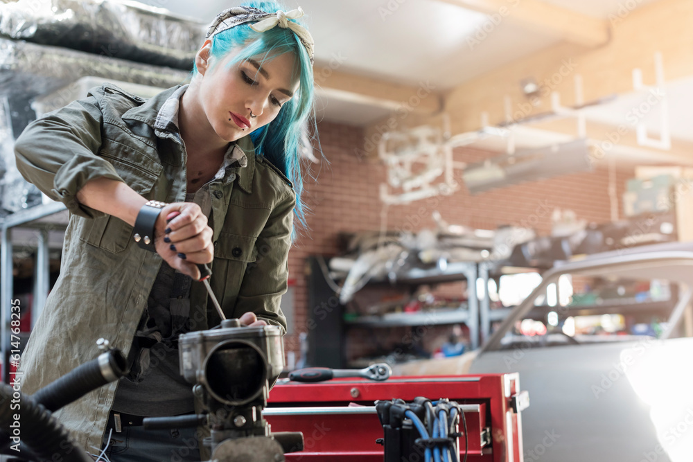 Young female mechanic blue hair repairing part in auto repair shop - obrazy, fototapety, plakaty 
