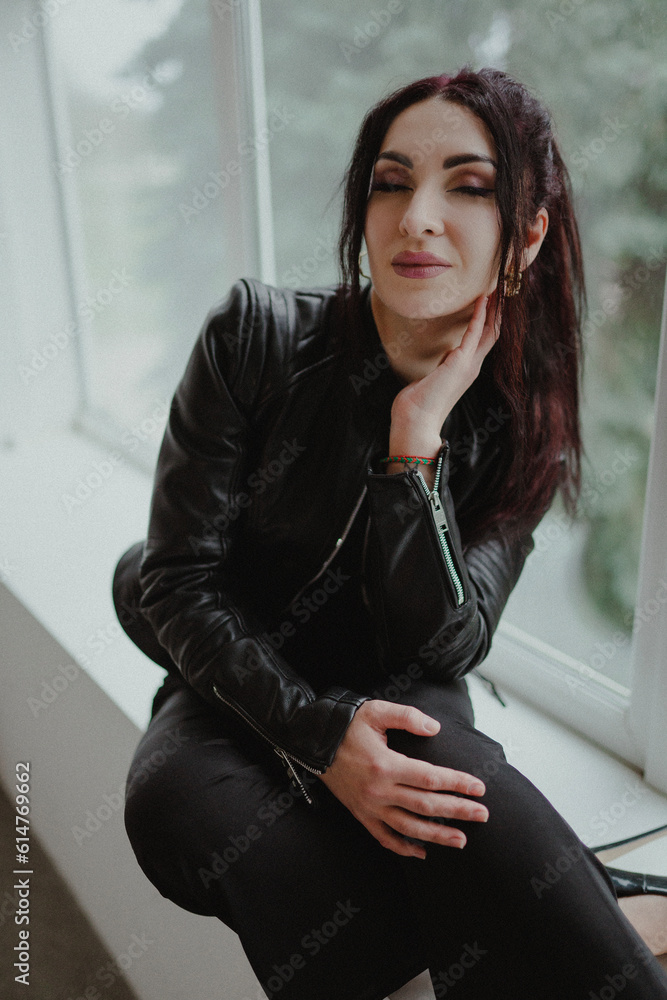 A hot brunette girl in a black dress and a black leather jacket sits on a window in a white studio and looks at the camera A hot brunette girl in a black dress and a black leather jacket sits on a window in a white studio and looks at the camera
