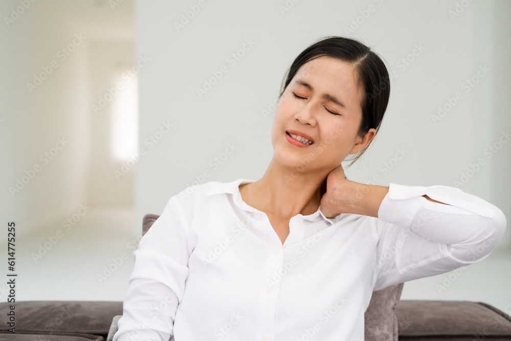 Unhealthy dark haired woman touching neck feeling pain and numbness, worried about muscle tension, osteochondrosis, wearing casual style shirt. Indoor living room isolated on white wall background. Unhealthy dark haired woman touching neck feeling pain and numbness, worried about muscle tension, osteochondrosis, wearing casual style shirt. Indoor living room isolated on white wall background.