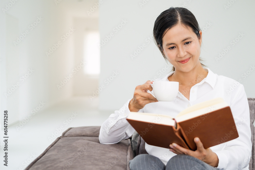 Young beautiful woman reading a book on sofa couch in living room at home . Asian female sitting on textile couch drinking coffee. White wall background, copy space, close up. Young beautiful woman reading a book on sofa couch in living room at home . Asian female sitting on textile couch drinking coffee. White wall background, copy space, close up.