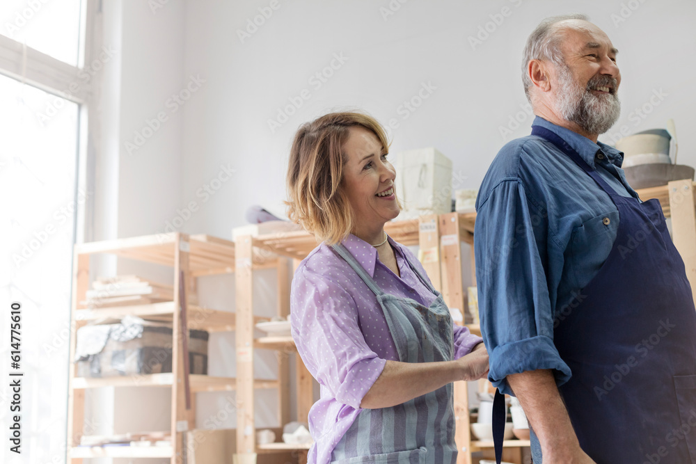 Fototapeta premium Smiling wife tying husband's apron in pottery studio