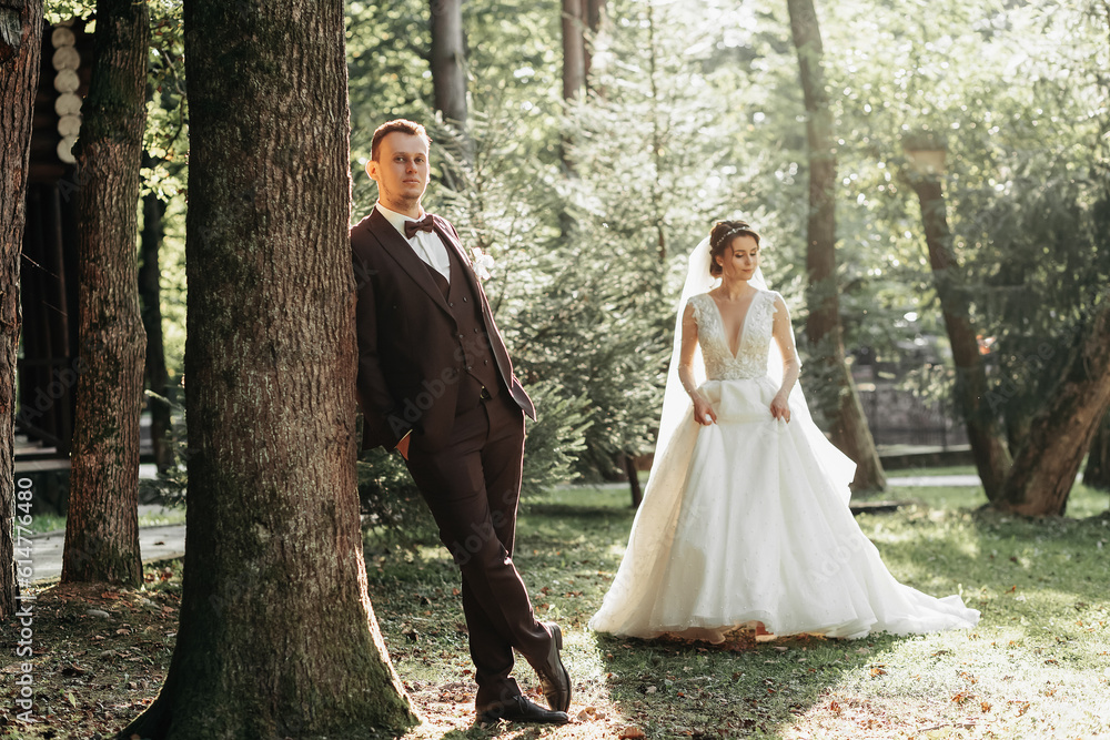 full-length portrait of a young bride and groom walking on a green golf course at sunset. Happy wedding couple, space for text. The groom leans against a tree, the bride is behind