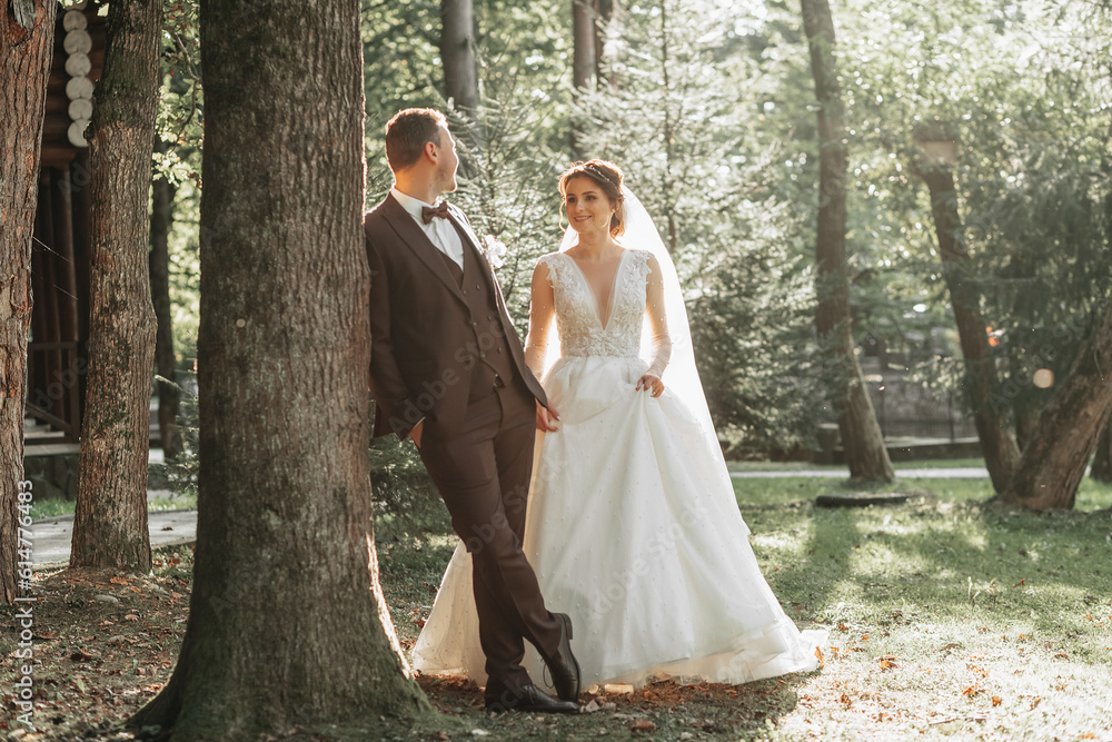full-length portrait of a young bride and groom walking on a green golf course at sunset. Happy wedding couple, space for text. The groom leans against a tree, the bride is behind full-length portrait of a young bride and groom walking on a green golf course at sunset. Happy wedding couple, space for text. The groom leans against a tree, the bride is behind