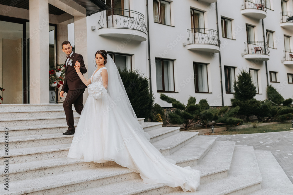 Fototapeta premium Groom and bride walking outdoors near a posh hotel. A long train of dress on the steps