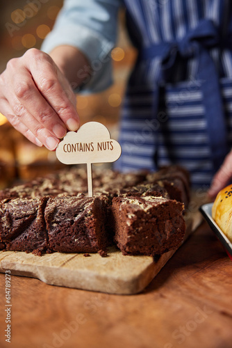 Sales Assistant In Bakery Putting Contains Nuts Label Into Freshly Baked Chocolate Brownies