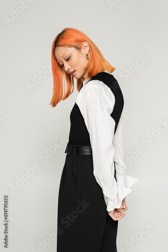 side view of expressive and young asian woman posing with hands behind back on grey background, dyed red hair, white shirt, vest and pants, business casual fashion, modern lifestyle
