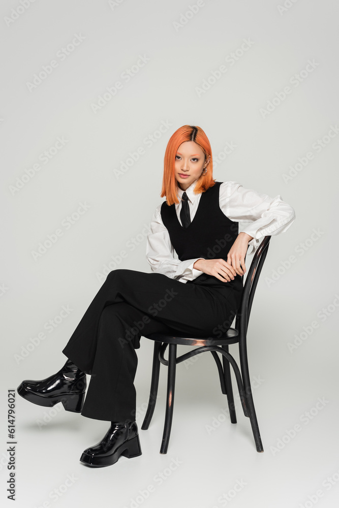 attractive and self-assured asian woman with colored red hair, in white shirt, black vest and pants sitting on chair and looking at camera on grey background, fashion shoot, full length attractive and self-assured asian woman with colored red hair, in white shirt, black vest and pants sitting on chair and looking at camera on grey background, fashion shoot, full length