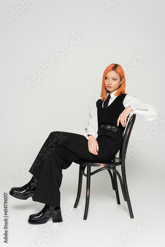 full length of redhead asian woman in black and white casual clothes posing on chair and looking away on grey background, colored red hair, shirt, tie, vest and pants, business casual style © LIGHTFIELD STUDIOS