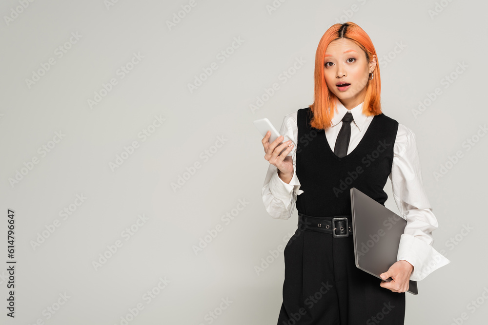 amazement, surprised asian woman with dyed red hair and open mouth standing with laptop and smartphone while looking at camera on grey background, white shirt, black vest and pants, business casual amazement, surprised asian woman with dyed red hair and open mouth standing with laptop and smartphone while looking at camera on grey background, white shirt, black vest and pants, business casual