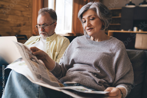 Elderly woman reading newspaper while man drowsing computer resting on couch together