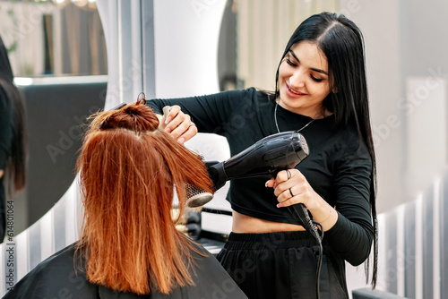 Beautiful woman stylist doing hair styling with hairdryer at client in modern beauty salon. Hairdresser makes hairstyle for middle-aged Caucasian woman with red hair.