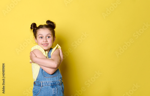 Caucasian happy emotional little kid girl, dressed in yellow t-shirt and blue denim jeans, hugging herself, smiling cutely, expressing positive emotions, isolated on yellow background with copy space