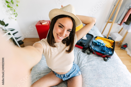 Beautiful young woman traveler taking selfie portrait with phone while preparing summer vacation suitcase at home. Smiling teenage girl posing for self portrait sitting on bed before going on holidays