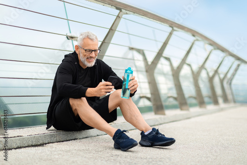 Mature sportsman having break, drinking water, using smartphone