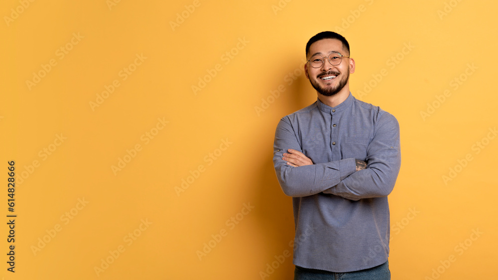 Confident young asian man standing with folded arms over yellow background - obrazy, fototapety, plakaty 