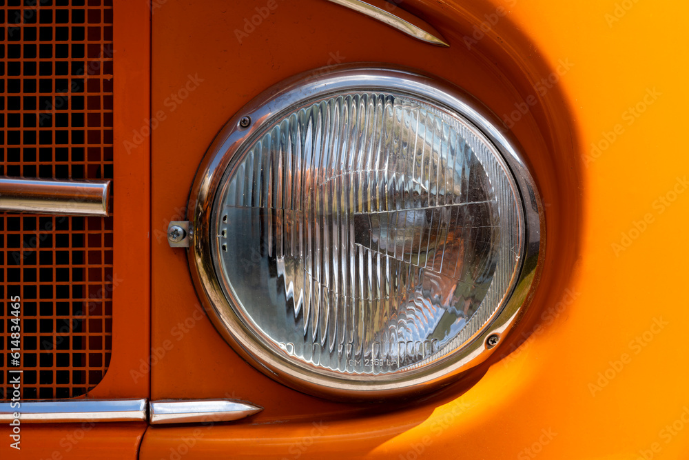 Headlight of a historic Oldtimer truck in bright sunlight. Orange livery with intense colorful gradient. Chrome parts, screws and paintwork surface of german vintage vehicle. Macro close up details.