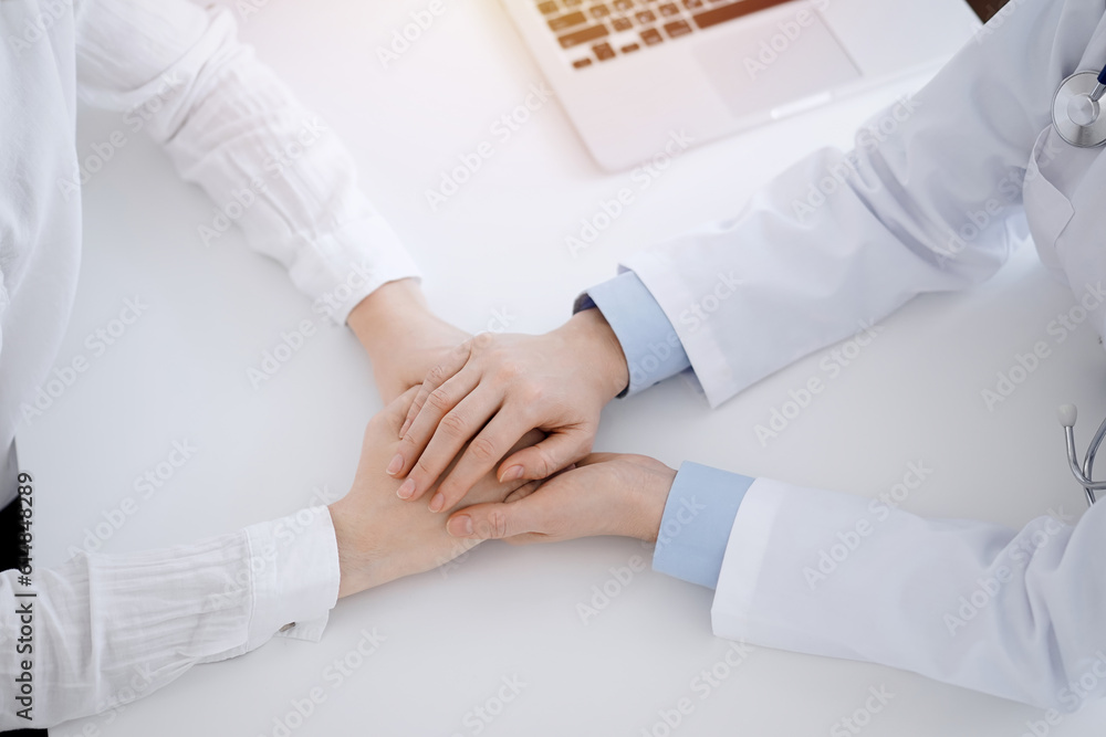 Fototapeta premium Doctor and patient sitting opposite each other at the table in clinic office. The focus is on female physician's hands reassuring woman, only hands, close up. Medicine concept