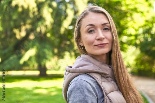 Radiant middle-aged Caucasian woman, smiling in the morning sunlight at the park, embracing the beauty of nature and finding joy in peaceful serenity © Asier