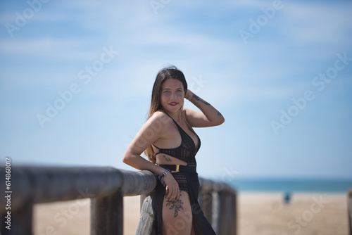 Attractive young woman with blue eyes, wearing black swimsuit and sarong, relaxed, solitary and calm, leaning on a railing of a wooden bridge, on the beach. Concept tranquility, peace, travel.