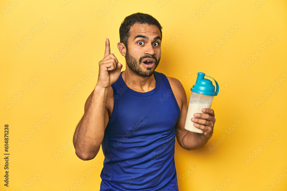 Athletic young Latino man holding a protein shake against a yellow background having an idea, inspiration concept. Athletic young Latino man holding a protein shake against a yellow background having an idea, inspiration concept.
