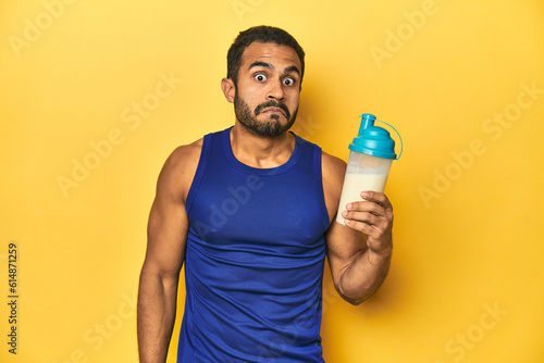 Obraz na plátně Athletic young Latino man holding a protein shake against a yellow background shrugs shoulders and open eyes confused