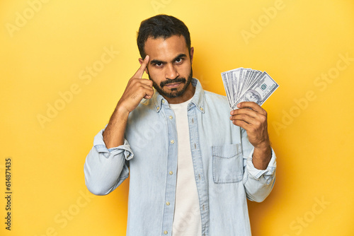 Young Latino man holding a bundle of dollars, yellow studio background, pointing temple with finger, thinking, focused on a task. photo