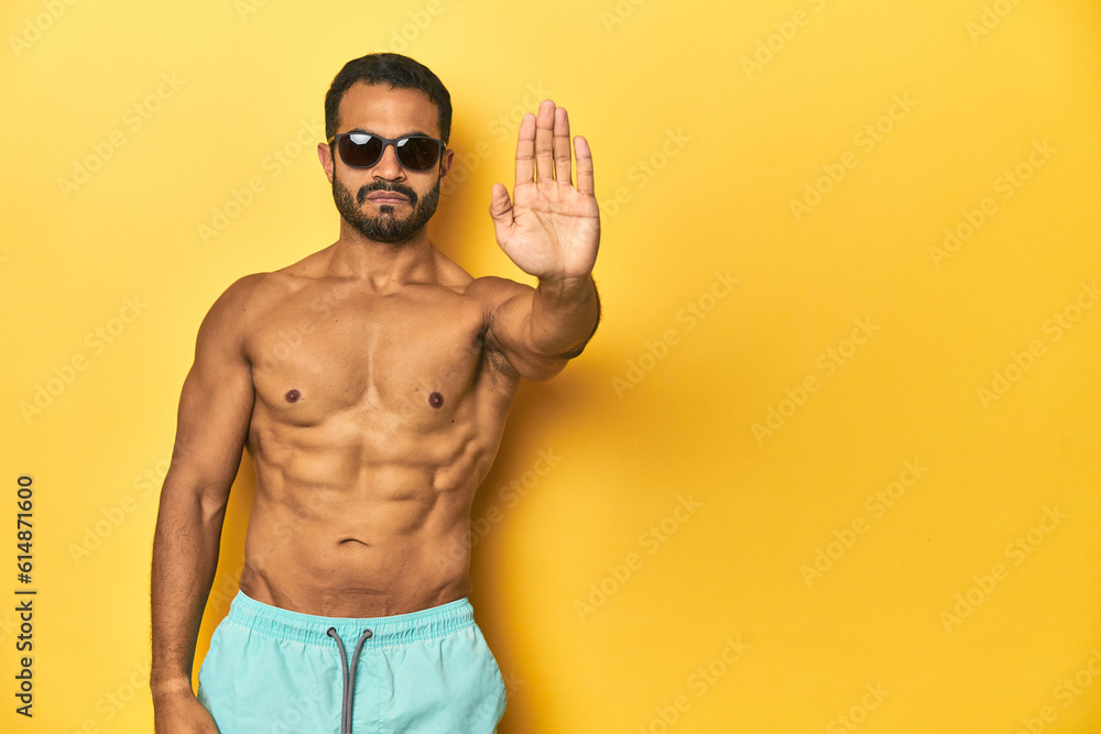 Fit young Latino man in swimwear and sunglasses, yellow studio background, standing with outstretched hand showing stop sign, preventing you. Fit young Latino man in swimwear and sunglasses, yellow studio background, standing with outstretched hand showing stop sign, preventing you.