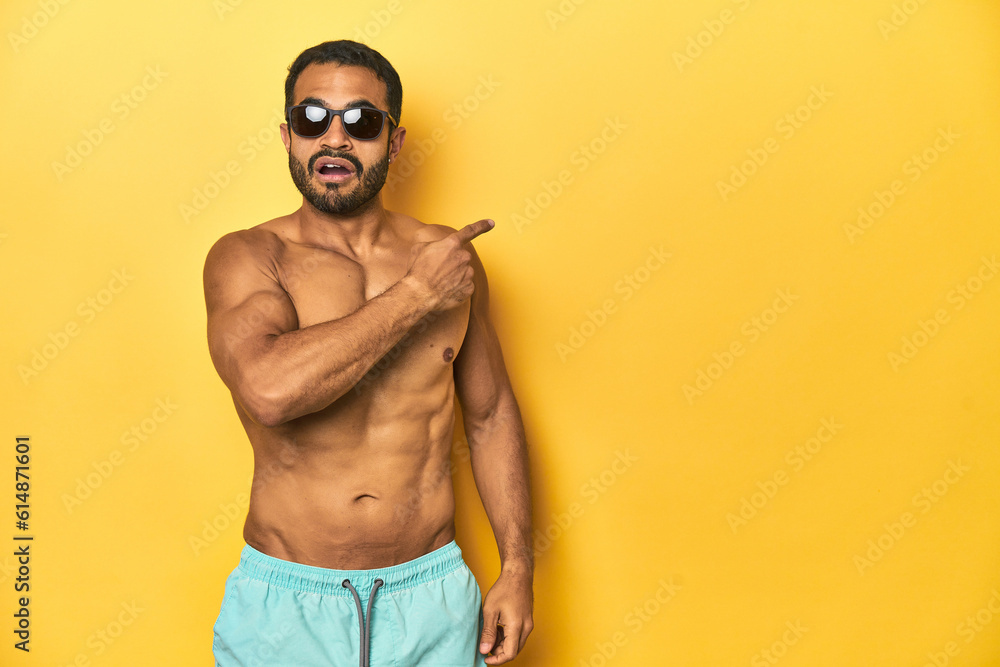 Fit young Latino man in swimwear and sunglasses, yellow studio background, pointing to the side Fit young Latino man in swimwear and sunglasses, yellow studio background, pointing to the side