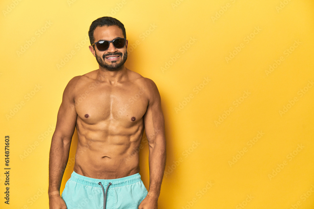 Fit young Latino man in swimwear and sunglasses, yellow studio background, happy, smiling and cheerful. Fit young Latino man in swimwear and sunglasses, yellow studio background, happy, smiling and cheerful.