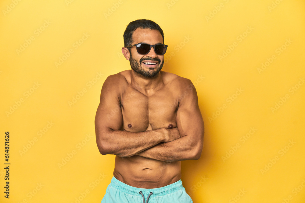 Fit young Latino man in swimwear and sunglasses, yellow studio background, laughing and having fun. Fit young Latino man in swimwear and sunglasses, yellow studio background, laughing and having fun.