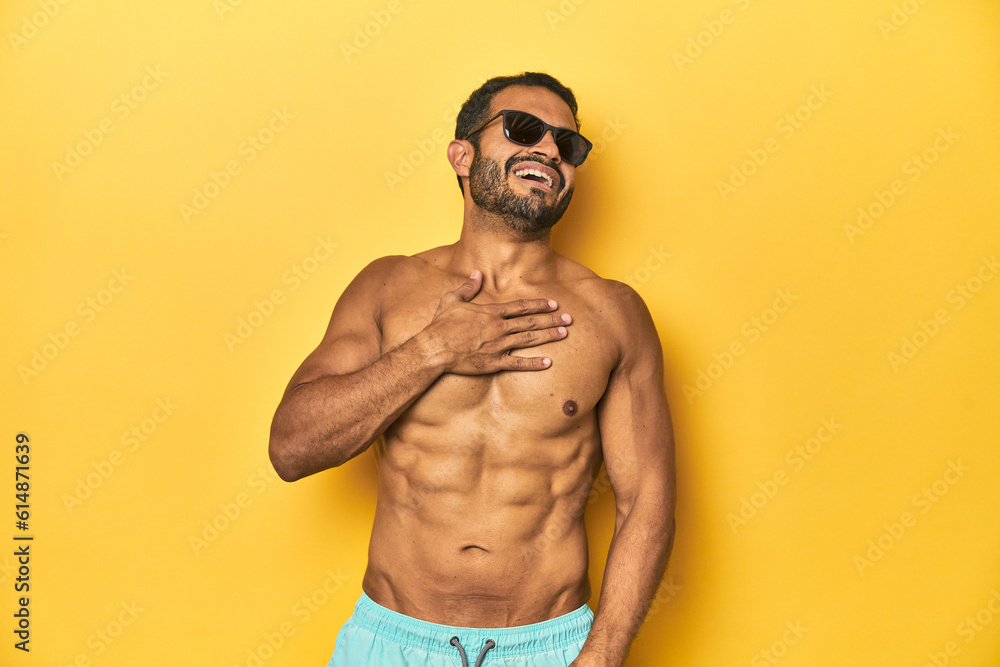 Fit young Latino man in swimwear and sunglasses, yellow studio background, laughs out loudly keeping hand on chest.