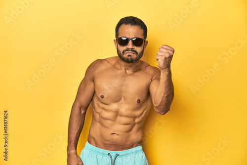 Fit young Latino man in swimwear and sunglasses, yellow studio background, showing fist to camera, aggressive facial expression.