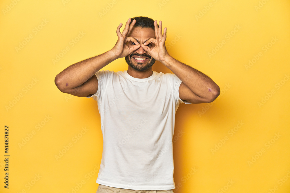 Casual young Latino man against a vibrant yellow studio background, showing okay sign over eyes Casual young Latino man against a vibrant yellow studio background, showing okay sign over eyes