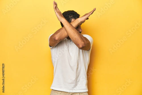 Tableau sur toile Casual young Latino man against a vibrant yellow studio background, keeping two arms crossed, denial concept