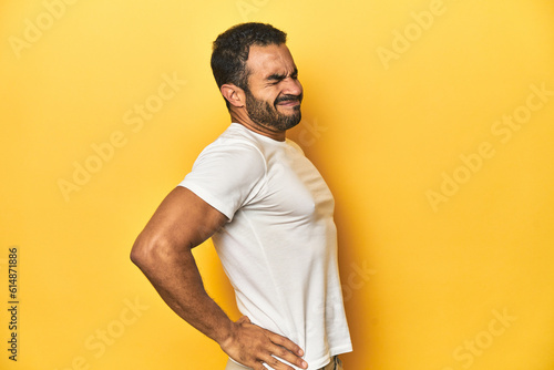 Casual young Latino man against a vibrant yellow studio background, suffering a back pain.