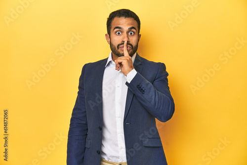 Professional young Latino man in business suit, yellow studio background, keeping a secret or asking for silence.