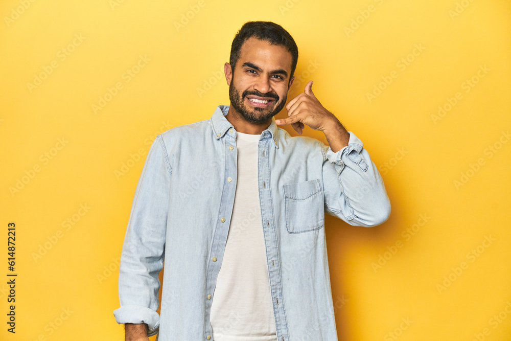 Casual young Latino man against a vibrant yellow studio background, showing a mobile phone call gesture with fingers.
