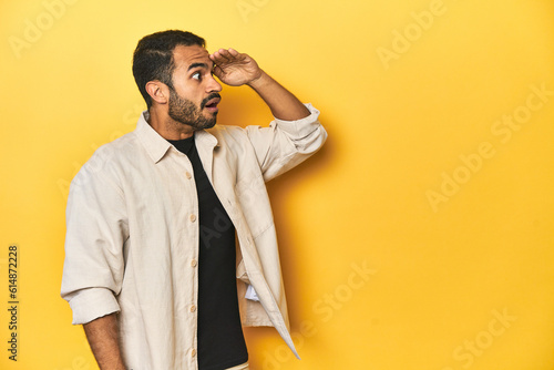 Casual young Latino man against a vibrant yellow studio background, looking far away keeping hand on forehead.