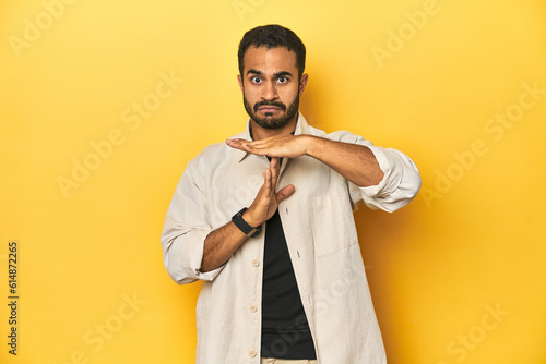 Casual young Latino man against a vibrant yellow studio background, showing a timeout gesture.